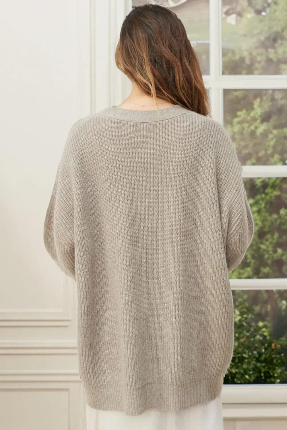 Person wearing a beige sweater standing in front of a window with greenery outside.