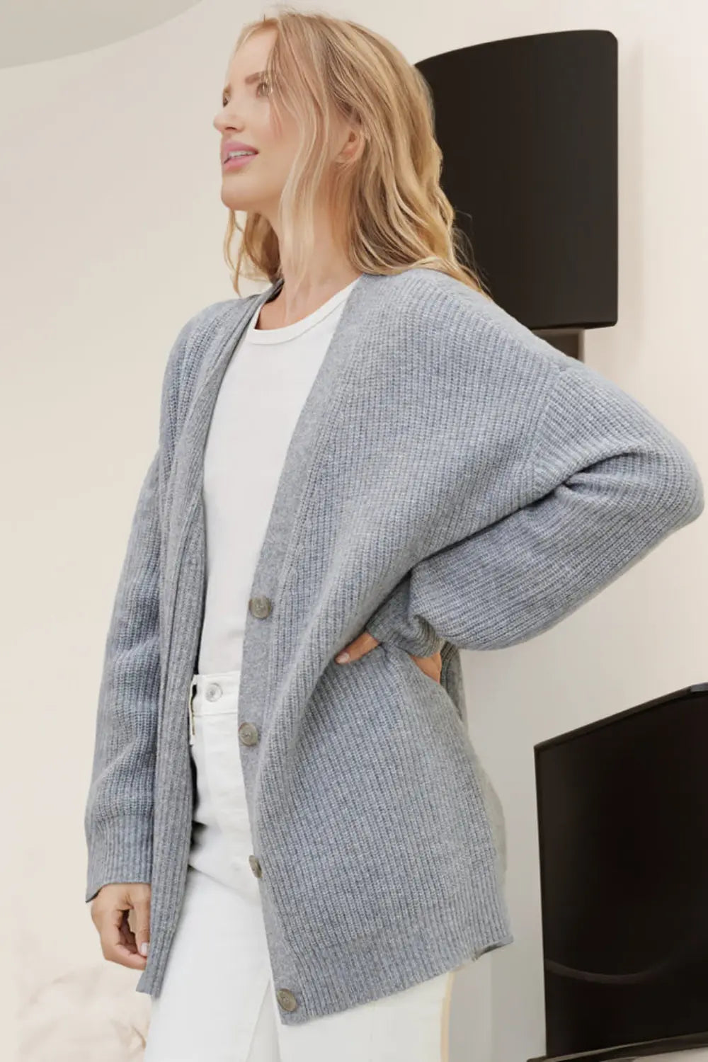 Woman wearing a gray cardigan over a white top and pants, standing indoors.