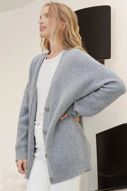 Woman wearing a gray cardigan over a white top and pants, standing indoors.